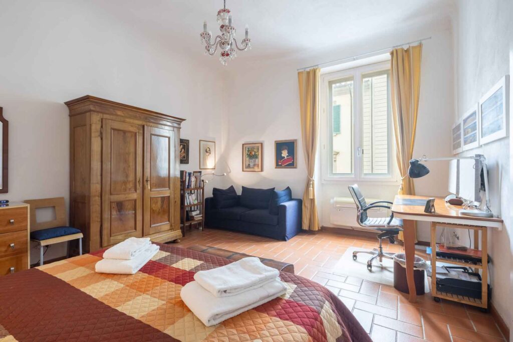 View across the spacious second living area of the Firenze Casa Florence Apartment, with its sofa, terracotta floor and wooden ceiling, looking through the adjoining terrace room towards the private green terrace.