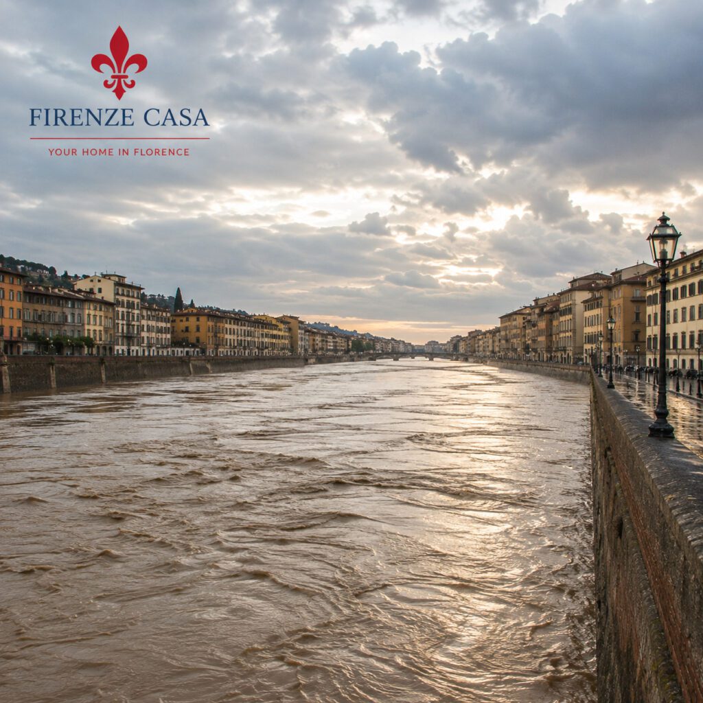 The Arno River flowing at flood level through Florence beneath dramatic storm clouds, illustrating the powerful force that has shaped the city's history for centuries.